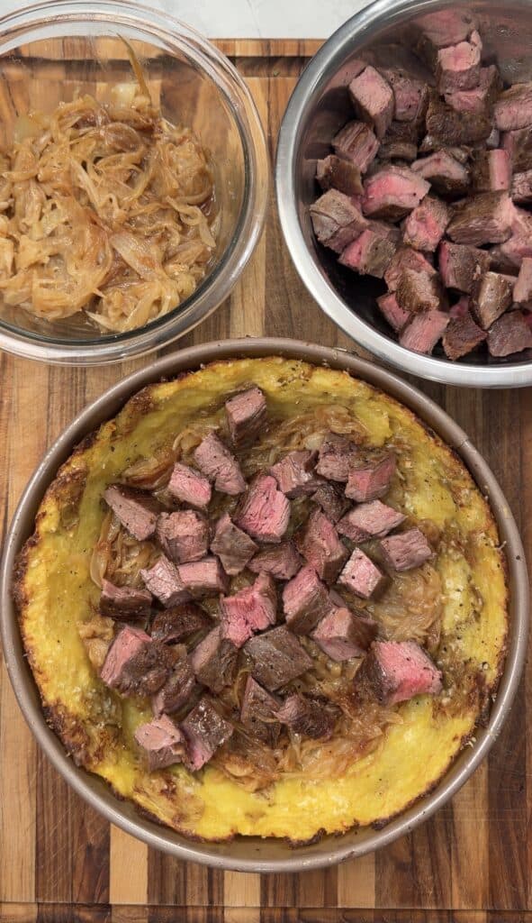 A cooked steak and onion tart on a wooden board, topped with cubed steak, evokes the hearty flavors of a classic Steak & Potato Pie. Two bowls nearby contain caramelized onions and more cubed steak.