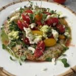 A colorful plate of grilled chicken paillard topped with a fresh salad with cherry tomatoes, microgreens, crumbled cheese, and a green dressing, served on a white plate with a bamboo-patterned rim. A whole tomato and a gold vase are in the background.