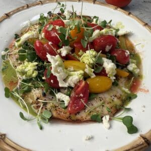 A colorful plate of grilled chicken paillard topped with a fresh salad with cherry tomatoes, microgreens, crumbled cheese, and a green dressing, served on a white plate with a bamboo-patterned rim. A whole tomato and a gold vase are in the background.