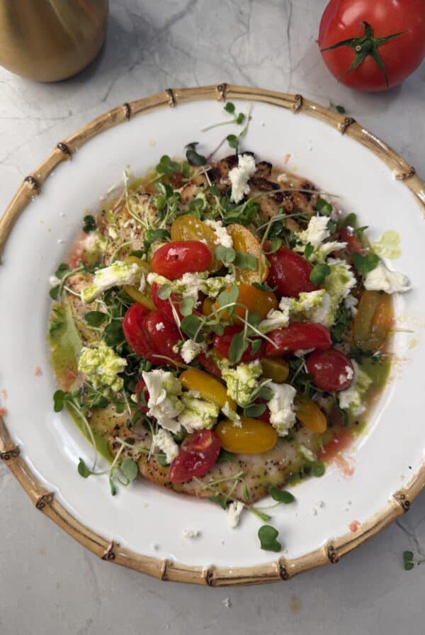 A white plate with grilled chicken paillard, colorful cherry tomatoes, crumbled cheese, microgreens, and a green herb sauce, on a marble surface with a whole tomato nearby.
