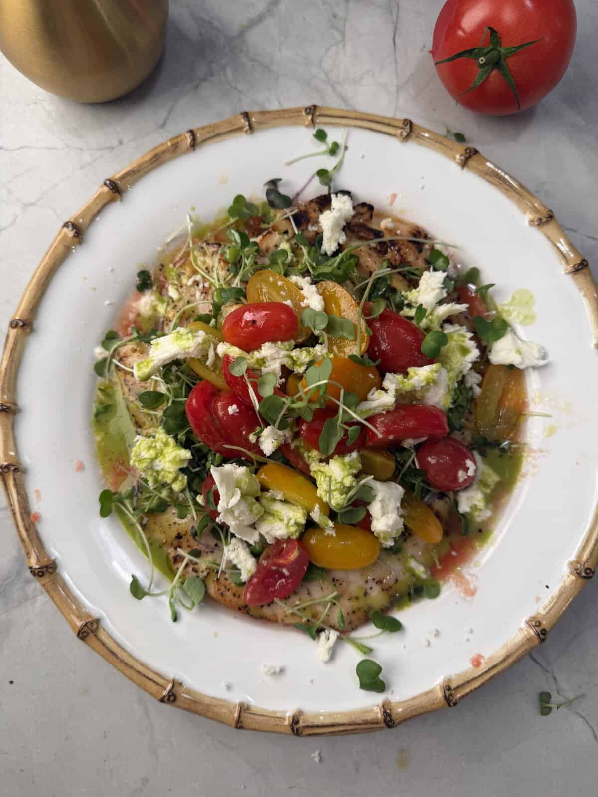 A white plate with grilled chicken, colorful cherry tomatoes, crumbled cheese, microgreens, and a green herb sauce, on a marble surface with a whole tomato nearby.