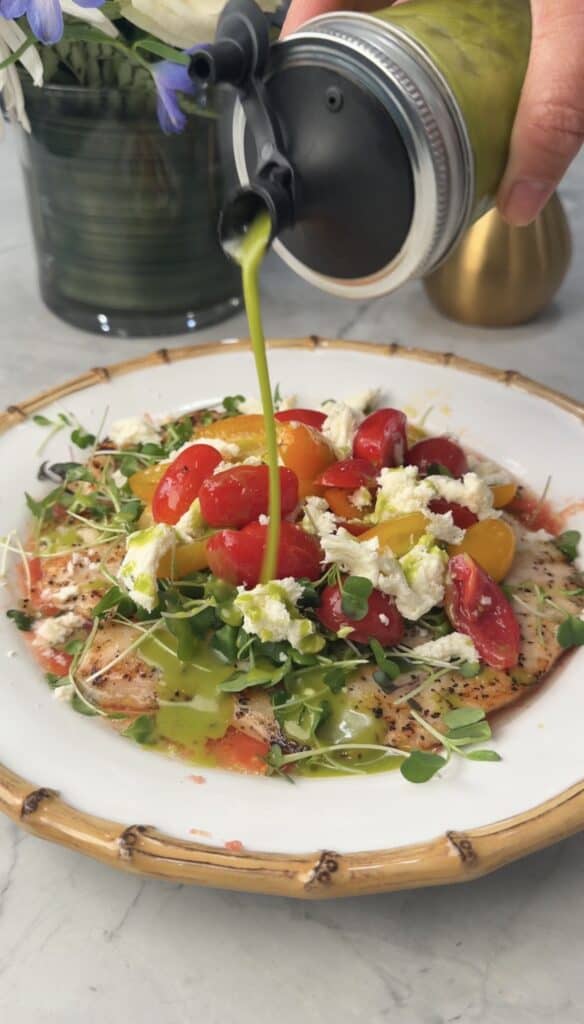 A hand pours green dressing over a plate of grilled chicken paillard topped with cherry tomatoes, microgreens, and crumbled cheese on a white plate with a bamboo rim.