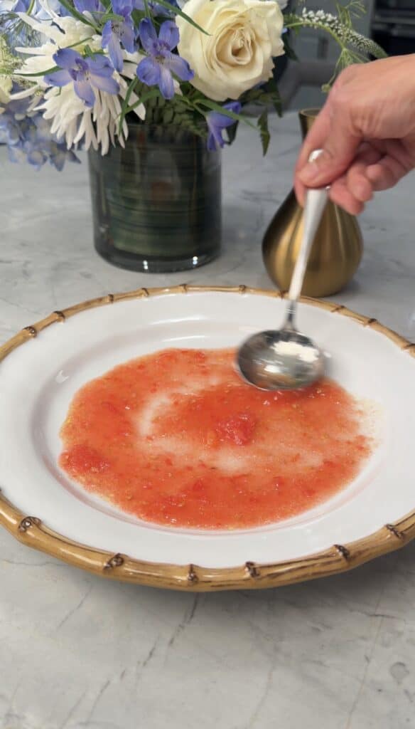 A hand holds a spoon over a bowl of tomato soup with foam on top, set on a marble table with flowers and a brass vase in the background.