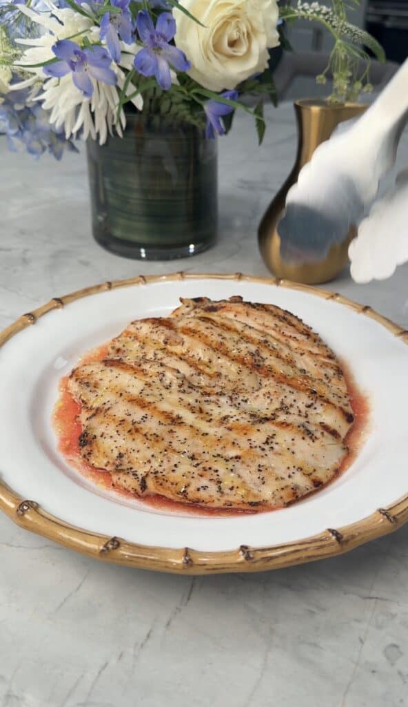Grilled chicken breast with grill marks on a white plate with a bamboo-style rim, with a floral arrangement and vase in the background.