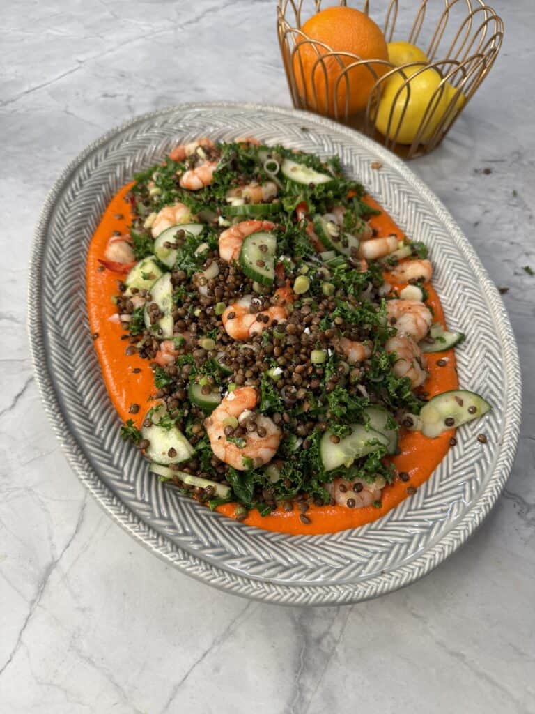 A silver platter with a crispy lentil salad topped with shrimp, sliced cucumber, crispy lentil salad, and herbs on a bed of red sauce. In the background, a wire basket holds an orange and two lemons on a marble surface.
