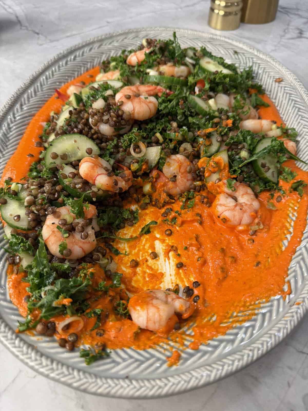 A platter of crispy lentil salad topped with shrimp salad with cucumbers, crispy lentil salad, herbs, and a bright orange sauce on a white oval dish, set on a marble surface.