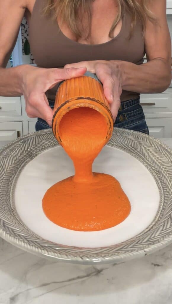A person pours thick, bright orange sauce from a blender onto a decorative plate in a kitchen, preparing to serve it alongside a crispy lentil salad.