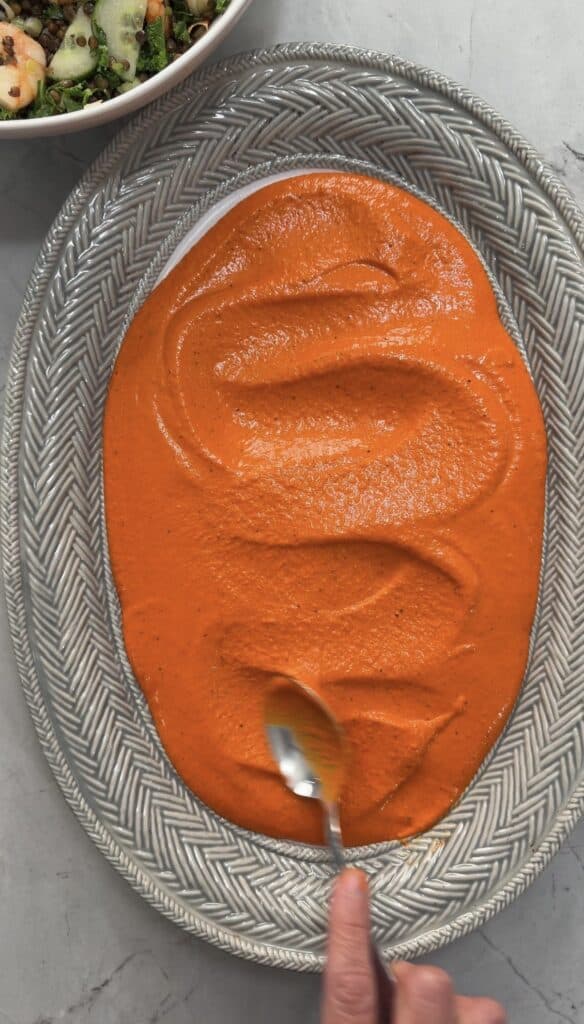 A hand spreads bright orange sauce with a spoon on an oval-patterned platter, while part of a crispy lentil salad bowl is visible in the top left corner.