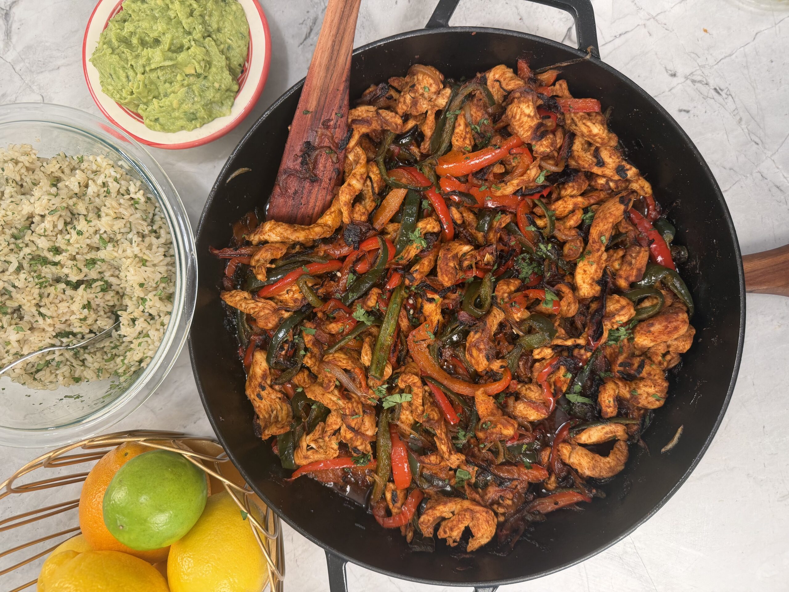 A pan filled with 30 minute skillet fajitas. The pan, filled with chicken, peppers, and onions sits beside a bowl of guacamole, a bowl of cilantro rice, and a basket with lemons and a lime on a marble countertop.