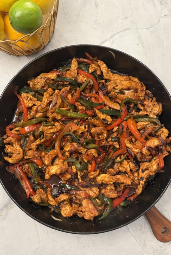 A skillet filled with cooked chicken strips, red and green bell peppers, and onions on a marble surface. A basket of lemons and a lime is in the corner.