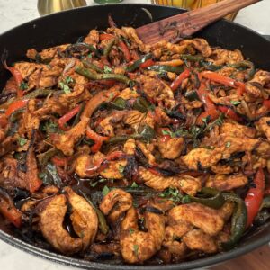 Colorful 30 minute fajitas with sautéed chicken, red and green peppers and onions in a black skillet, garnished with herbs. A wooden spatula rests in the skillet. Lemons, limes, and a pepper mill are visible in the background.