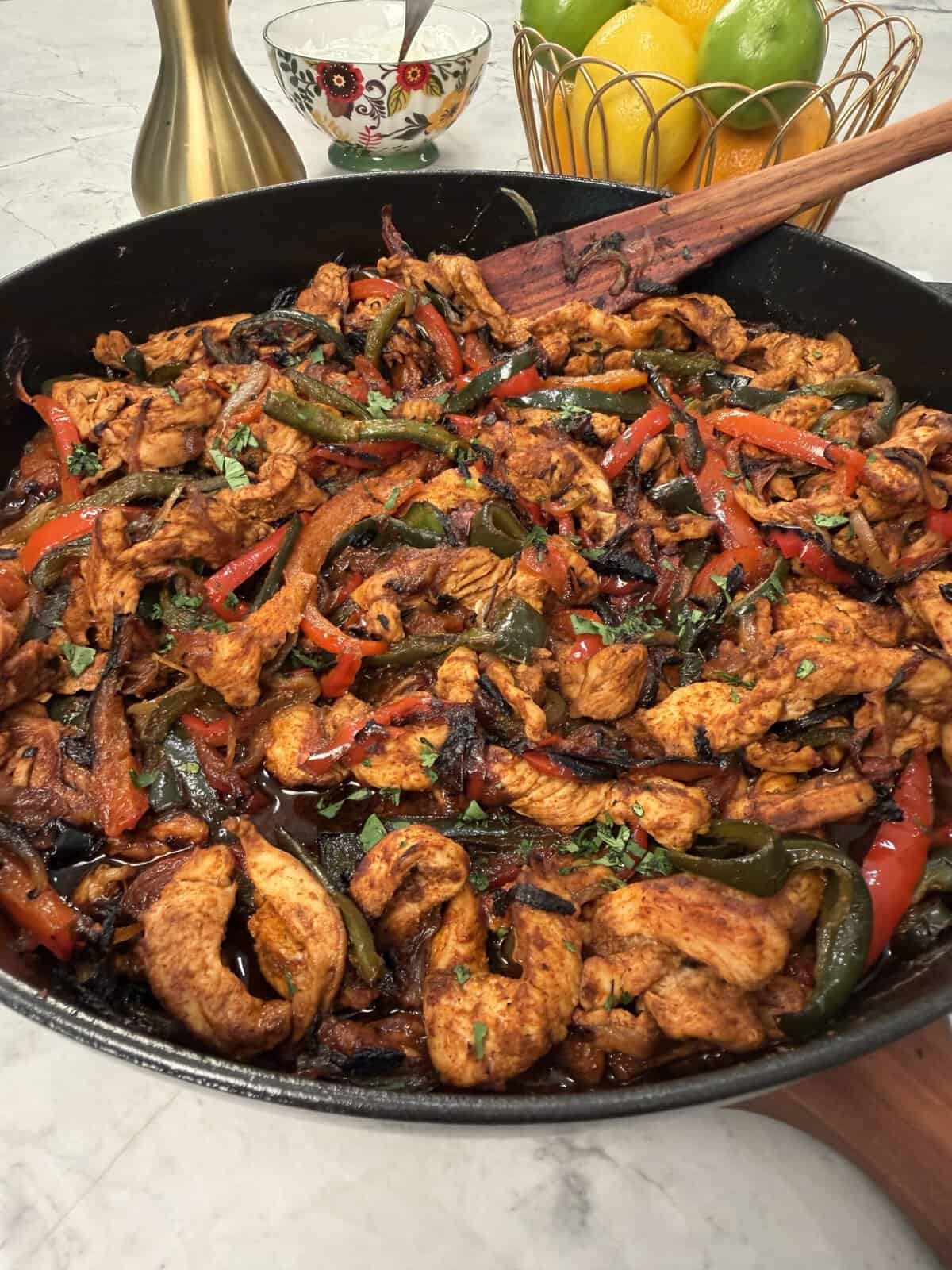 Colorful chicken fajitas with sautéed red and green peppers and onions in a black skillet, garnished with herbs. A wooden spatula rests in the skillet. Lemons, limes, and a pepper mill are visible in the background.