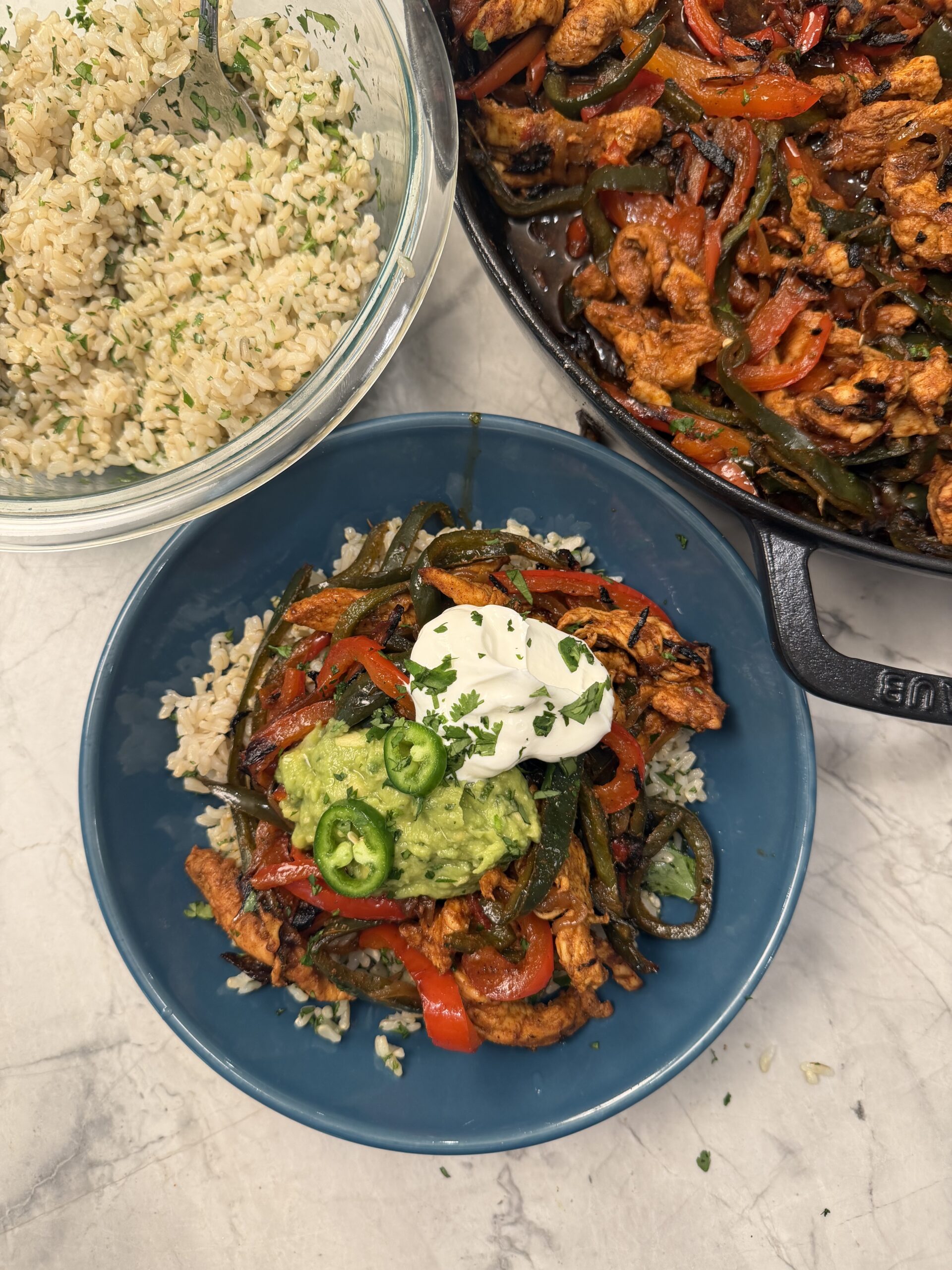 A blue bowl with rice topped with 30 minute skillet fajita chicken, sautéed peppers, guacamole, sour cream, and sliced jalapeños. Next to it are a bowl of rice and a skillet of fajita mixture on a marble surface.