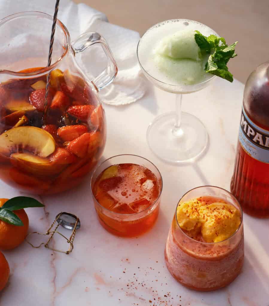 A pitcher of sangria with fruit, two cocktails in glasses, a frothy green drink garnished with mint, a Campari bottle, oranges, and a white napkin on a marble surface.