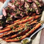 Grilled sliced steak and charred carrots topped with green herb sauce, served on a rectangular platter with a spoon of sauce on the side.