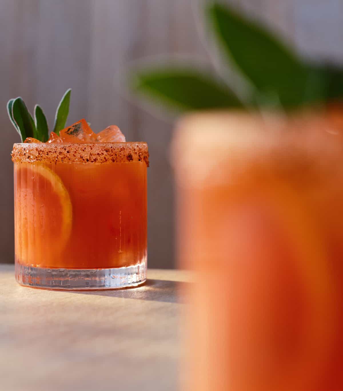 A glass of orange cocktail with ice, an orange slice, a spiced rim, and green herb garnish sits on a table with a blurred similar drink in the foreground.