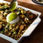 A spoon drizzles green sauce over burrata on a baked dish filled with roasted zucchini, corn, herbs, and scallions, in a white rectangular baking dish.