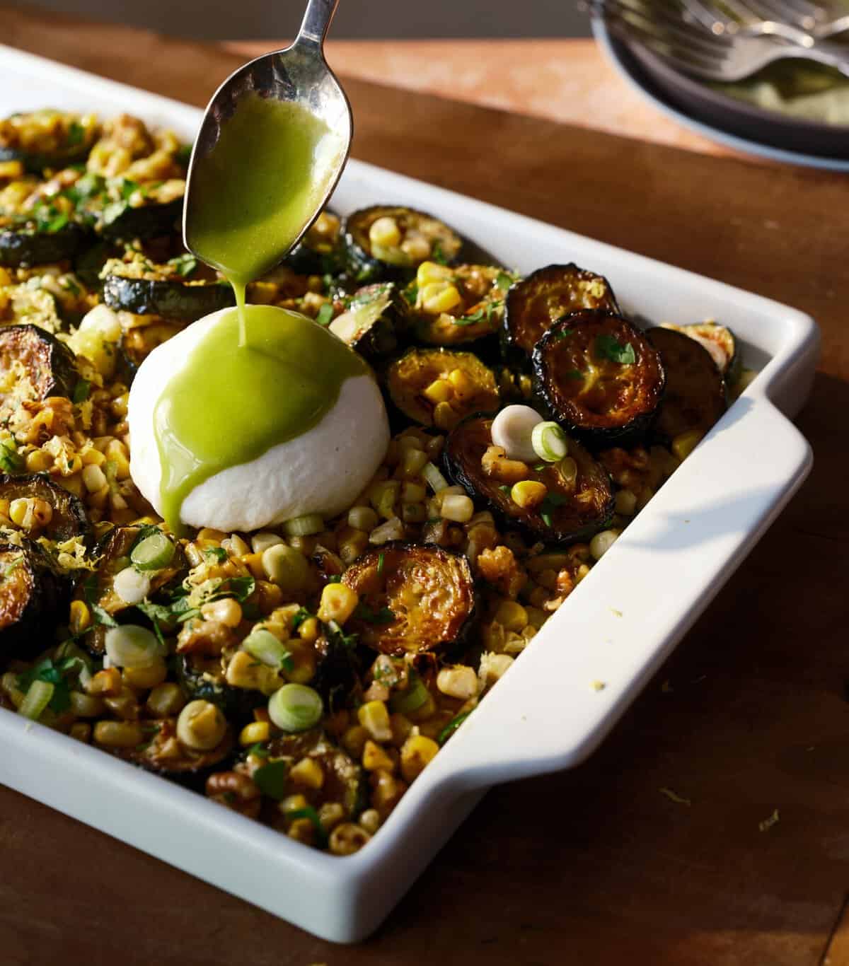 A spoon drizzles green sauce over burrata on a baked dish filled with roasted zucchini, corn, herbs, and scallions, in a white rectangular baking dish.