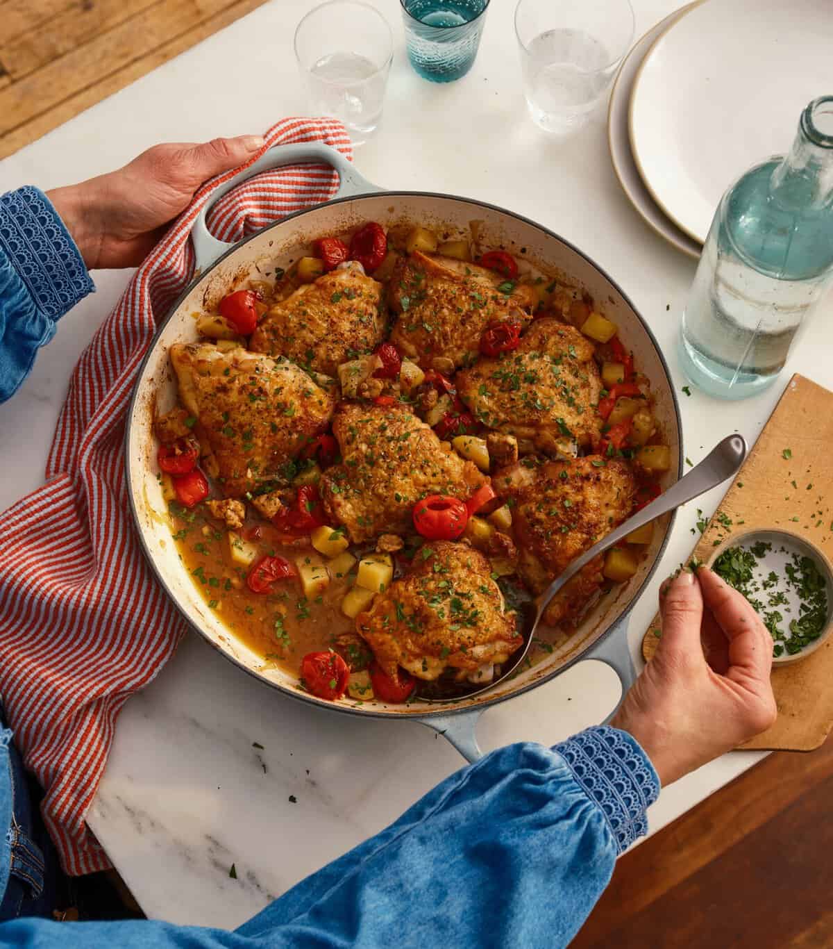 A person holds a skillet filled with cooked chicken thighs, potatoes, and tomatoes, garnished with herbs. The dish is on a white table beside drinking glasses, plates, a water bottle, and a red striped towel.