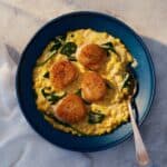 A blue bowl with four seared scallops on creamy corn and spinach, with a spoon resting inside. A white cloth is nearby on the textured surface.