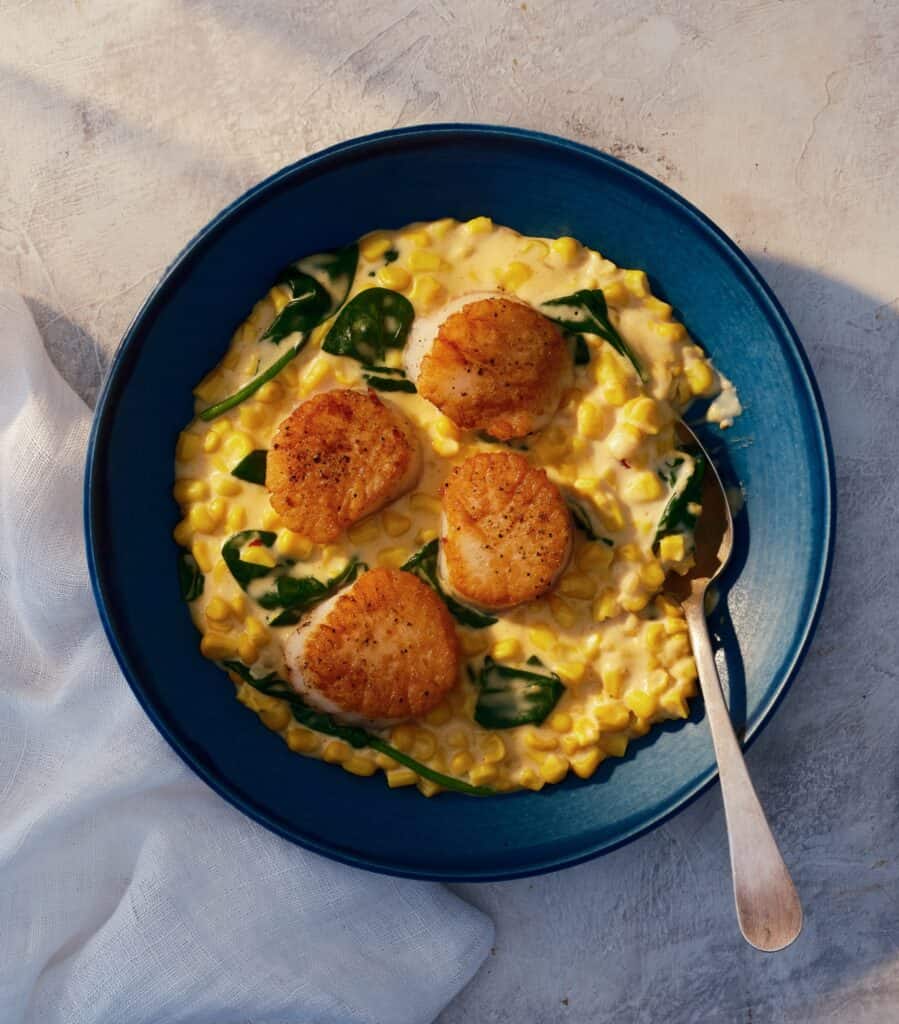 A blue bowl with four seared scallops on creamy corn and spinach, with a spoon resting inside. A white cloth is nearby on the textured surface.