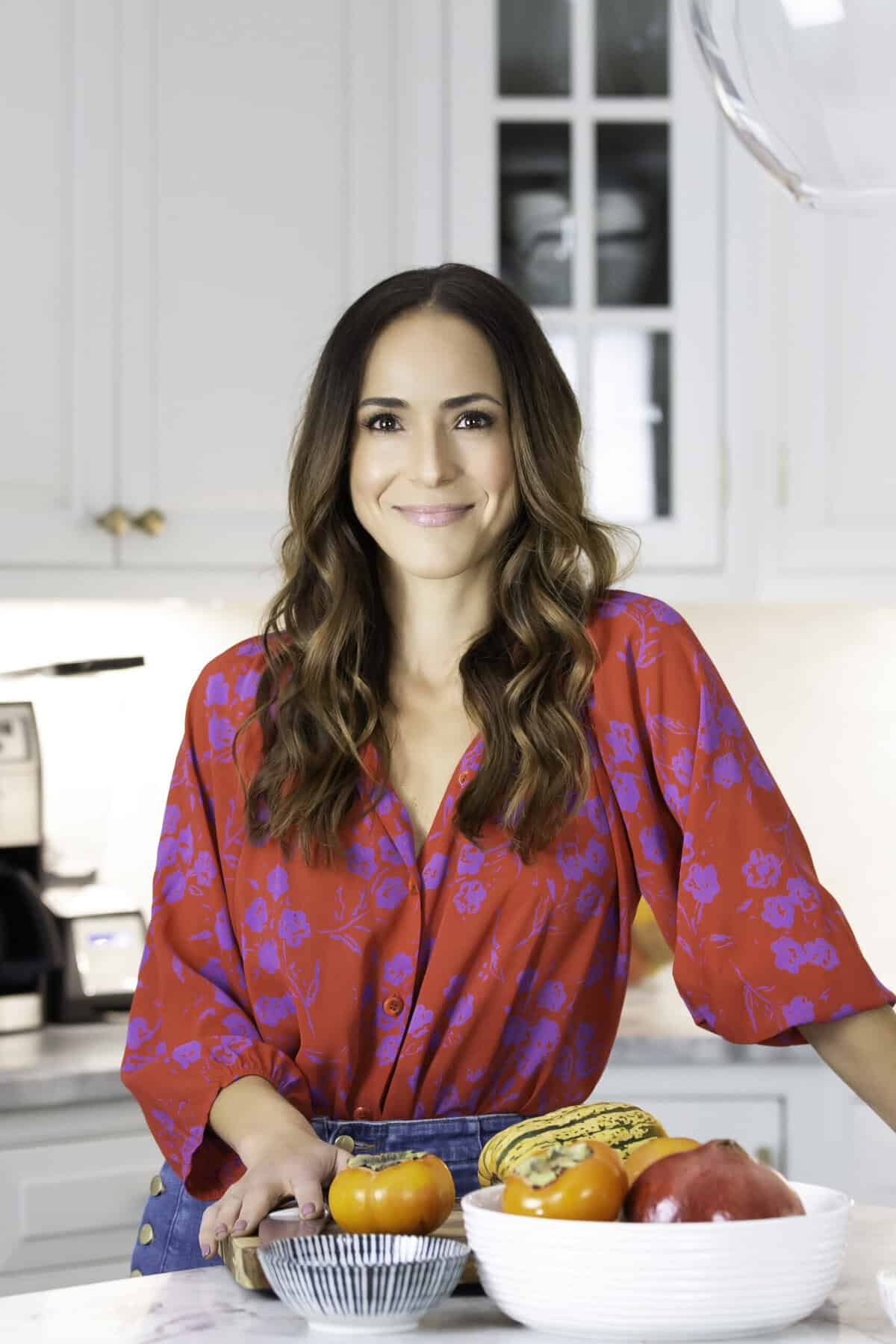 A woman with long brown hair in a red floral blouse stands in a bright kitchen, smiling, with a bowl of assorted fruits and vegetables on the counter in front of her.