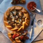 A rustic galette with roasted vegetables sits on parchment paper, surrounded by sliced pieces topped with tomato chutney. A jar of chutney, knife, and glass of water are nearby on a wooden surface.