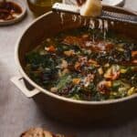A hand grates cheese over a pot of vegetable soup with greens and beans. Slices of bread and a small dish of chili oil are nearby on the table.