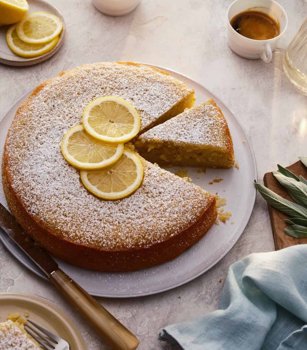 A round lemon cake dusted with powdered sugar, topped with lemon slices. One slice is cut. A knife, a cup of espresso, lemons, and a blue napkin are nearby on a light surface.