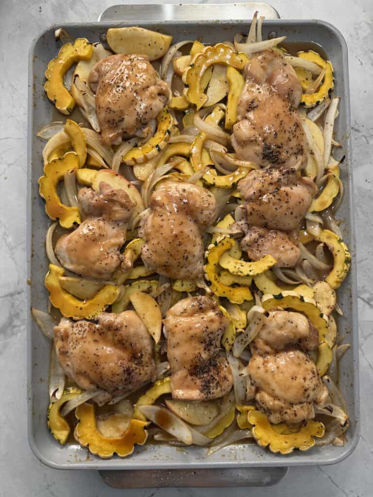 A baking sheet filled with maple dijon chicken thighs, sliced delicata squash, and onions, all arranged in rows and ready to be roasted in the oven.