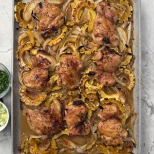 A sheet pan filled with roasted maple dijon chicken thighs, sliced delicata squash, and onions, garnished with herbs. Two small bowls with chopped green onions and herbs sit to the side.