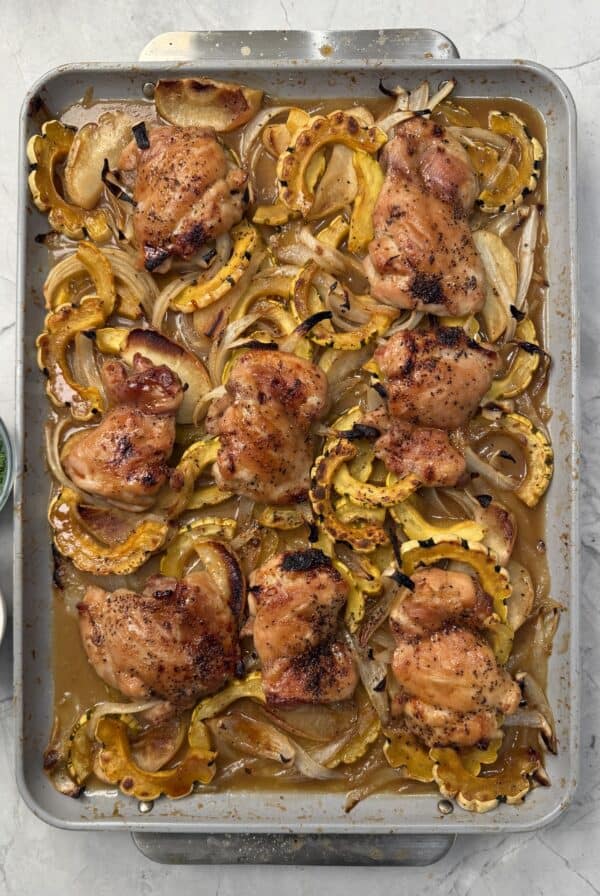 A sheet pan filled with roasted maple dijon chicken thighs, sliced delicata squash, and onions, garnished with herbs. Two small bowls with chopped green onions and herbs sit to the side.