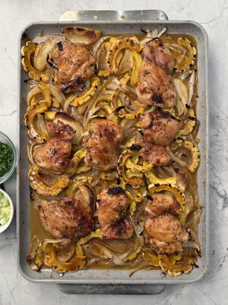 A sheet pan filled with roasted maple dijon chicken thighs, sliced delicata squash, and onions, garnished with herbs. Two small bowls with chopped green onions and herbs sit to the side.