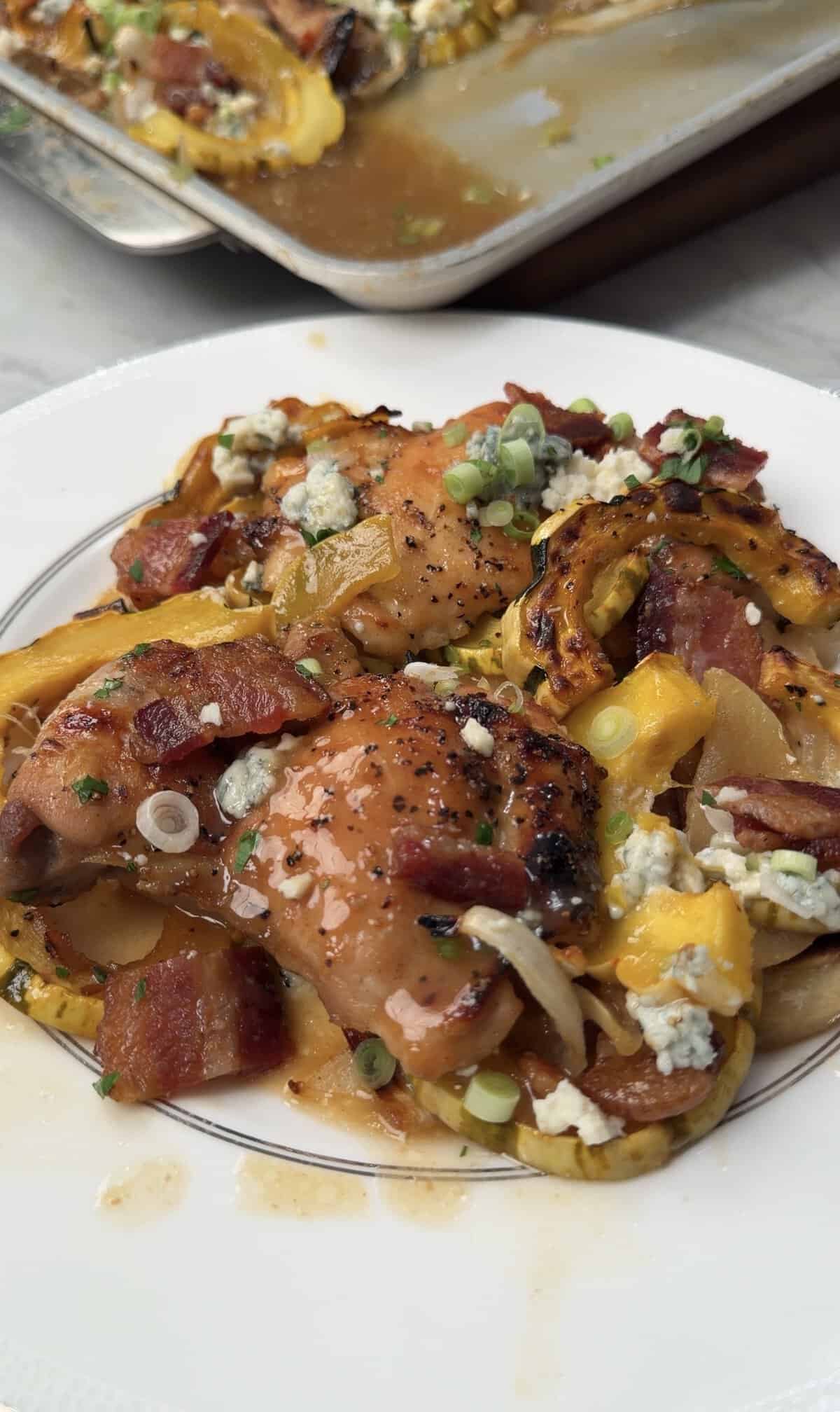 A plate of roasted chicken topped with bacon, blue cheese, green onions, and sliced squash, on a white dish with a pan of more food in the background.