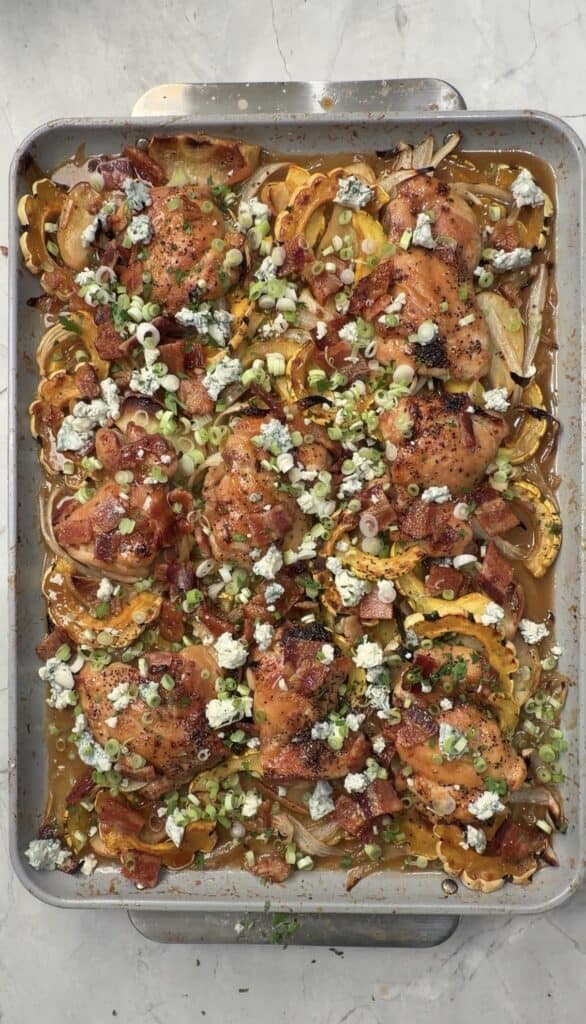 A baking sheet with roasted chicken thighs, sliced squash, onions, crumbled bacon, blue cheese, and chopped green onions on a marble countertop.
