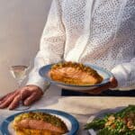 Person in a white eyelet blouse holding a plate of salmon with herbs and sauce, next to a glass of white wine and a plate of asparagus with walnuts.
