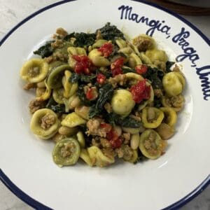 A plate of Sausage & Kale Pasta featuring orecchiette with savory sausage, tender kale, white beans, and vibrant red peppers, served on a white plate with a blue rim and Italian writing along the edge.