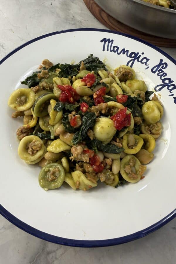 A plate of Sausage & Kale Pasta featuring orecchiette with savory sausage, tender kale, white beans, and vibrant red peppers, served on a white plate with a blue rim and Italian writing along the edge.