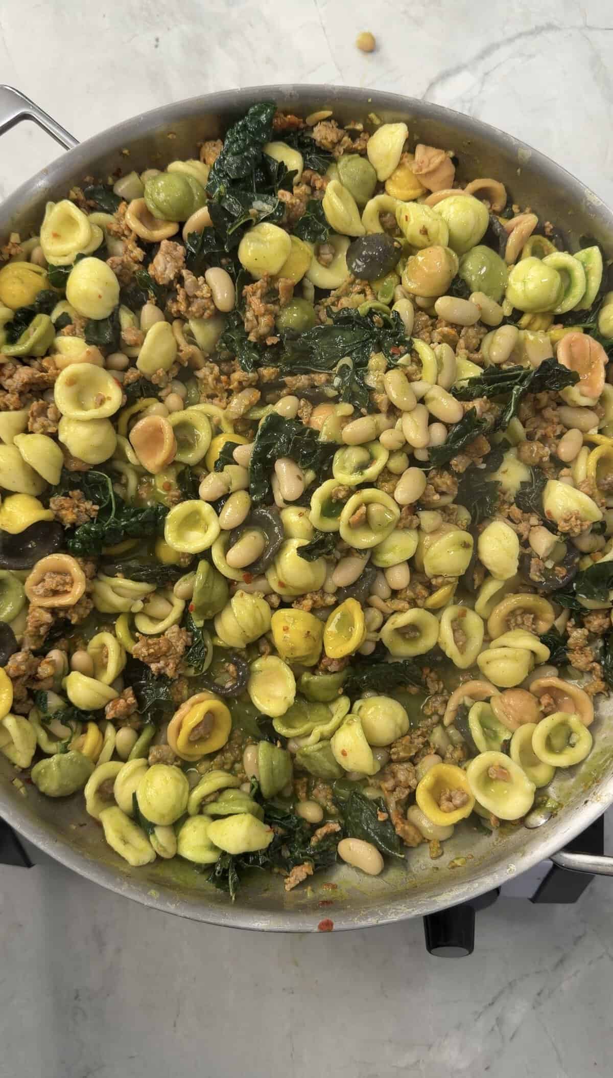 A pan filled with Sausage & Kale Pasta, combining orecchiette, savory ground meat, white beans, kale, and green vegetables, all mixed together.