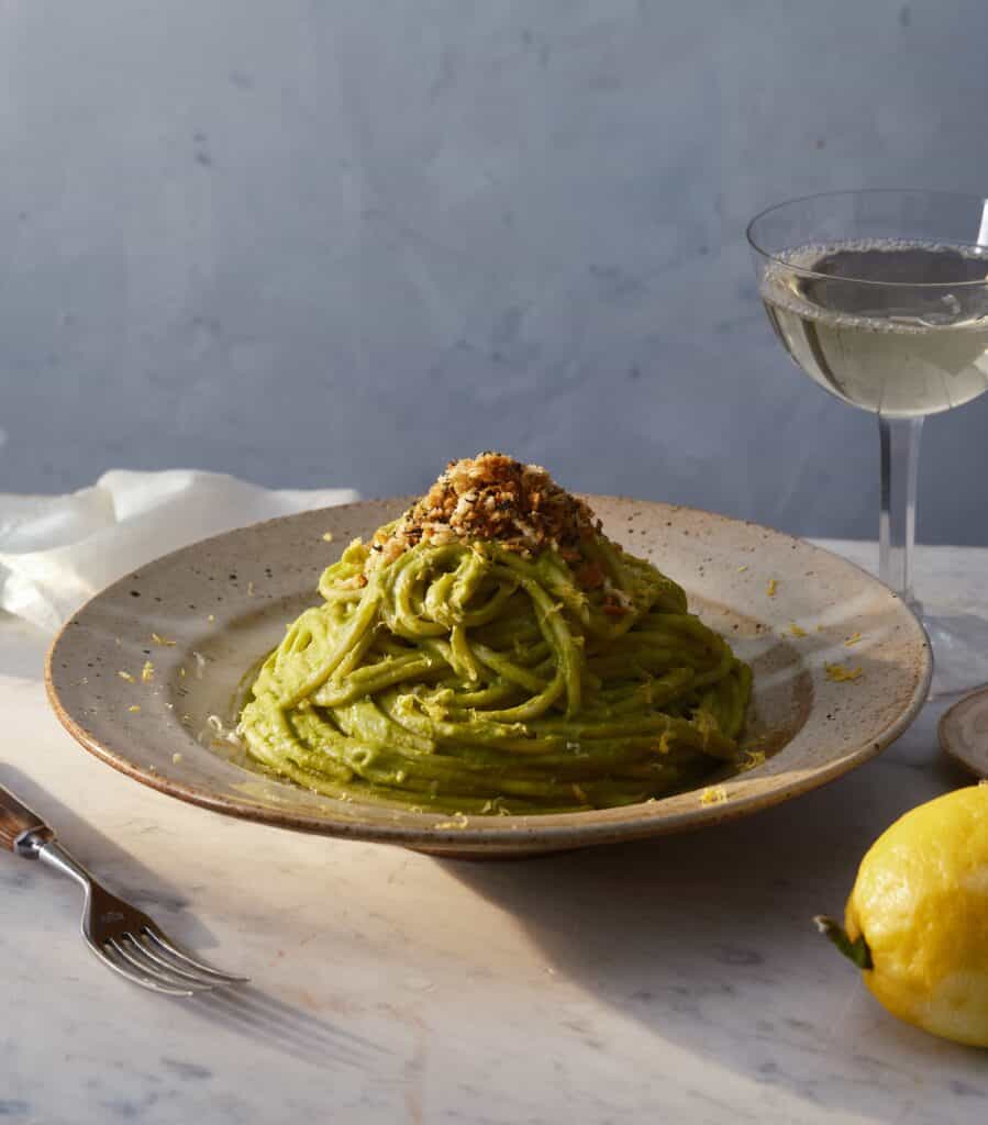A plate of green pasta topped with chopped nuts sits on a marble table beside a fork, a lemon, and a glass of white wine. The background is softly lit with neutral colors.