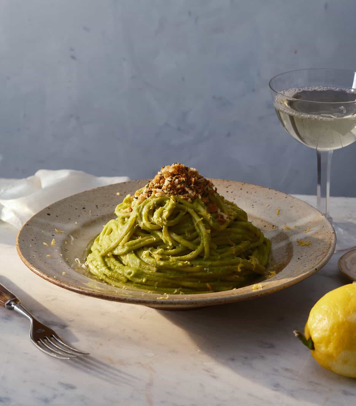 A plate of green pasta topped with chopped nuts sits on a marble table beside a fork, a lemon, and a glass of white wine. The background is softly lit with neutral colors.