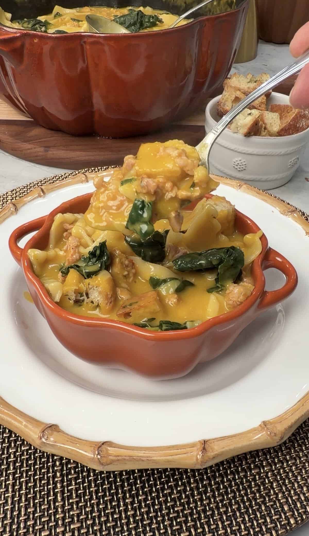 A spoonful of savory pumpkin lasagna soup with sausage and greens being lifted from a pumpkin-shaped bowl.