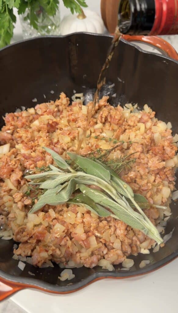 Wine being poured from a bottle into a cast iron pan of sautéed meat, onions, sage, and thyme for a savory pumpkin lasagna soup.
