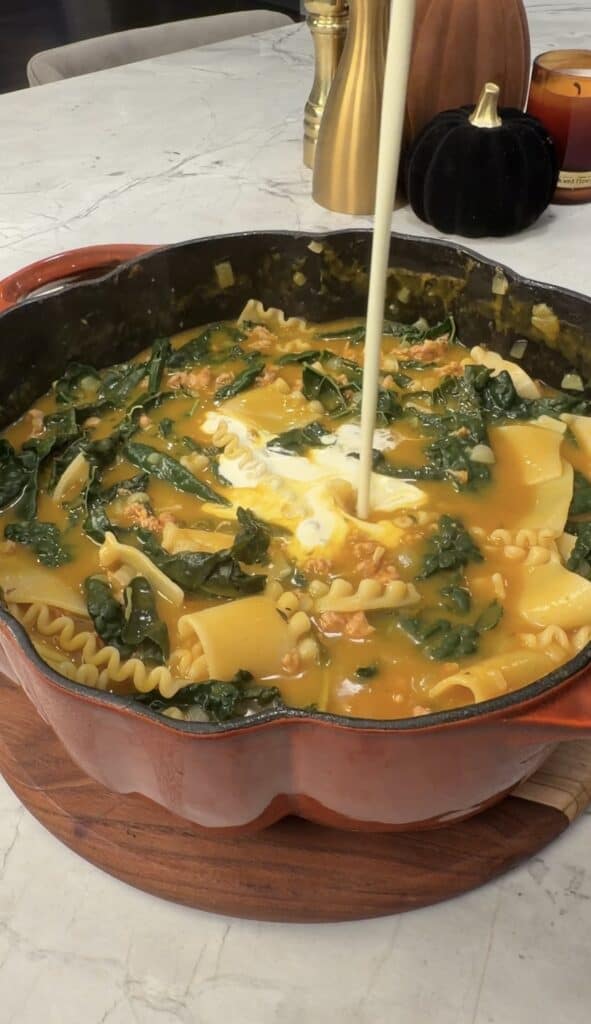 Cream being poured into a red Dutch oven filled with a savory pumpkin lasagna soup containing noodles and leafy greens.