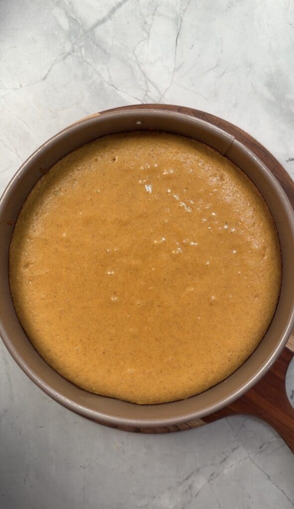 A round, orange pumpkin olive oil cake in a metal baking pan, resting on a wooden board.