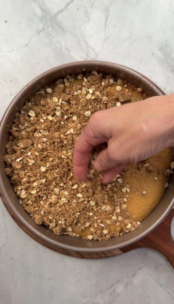 A hand sprinkles an oat crumble topping onto the filling of a pumpkin olive oil cake in a round cake pan.