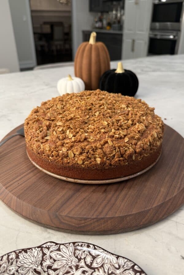 A round pumpkin olive oil cake with a thick oat streusel topping sits on a wooden board, with three decorative pumpkins in the background.