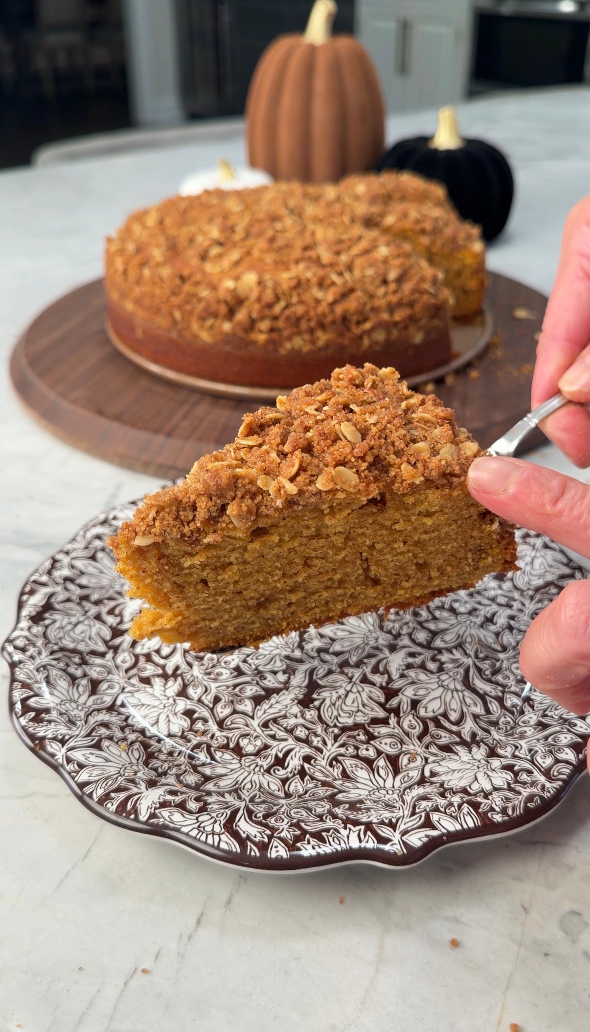 A hand uses a fork to lift a slice of pumpkin olive oil cake above a patterned plate, with the whole cake and decorative pumpkins in the background.