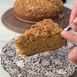 A hand holds a fork with a slice of pumpkin olive oil cake over a decorative floral plate, with the whole cake and decorative pumpkins in the background.
