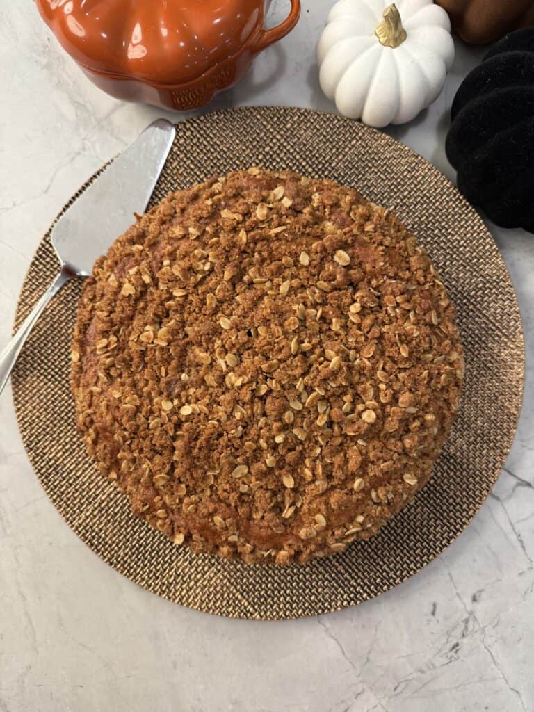 A round pumpkin olive oil cake topped with a generous layer of oat crumble sits on a woven placemat next to a metal cake server, with decorative pumpkins in the background.